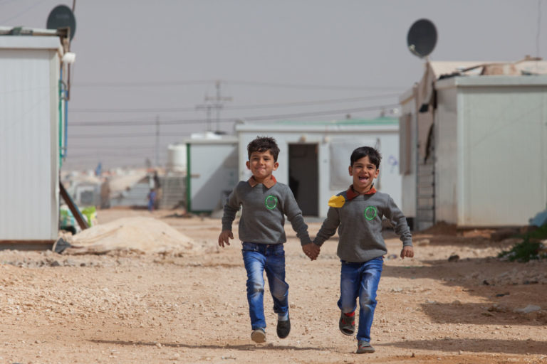 Five year old twins Besher and Ghanem running in the road between people's container homes. The family fled Syria and have lived in the camp in northern Jordan for a year and eight months. You can support children like Besher and Ghanem through a gift in your Will. Join the Unicef Generation.